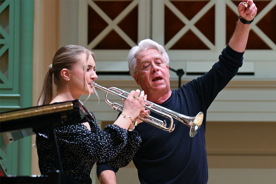 A man with light hair, wearing a black top, raising his hands up as he is teaching a music class, with a female trumpet player playing next to him.